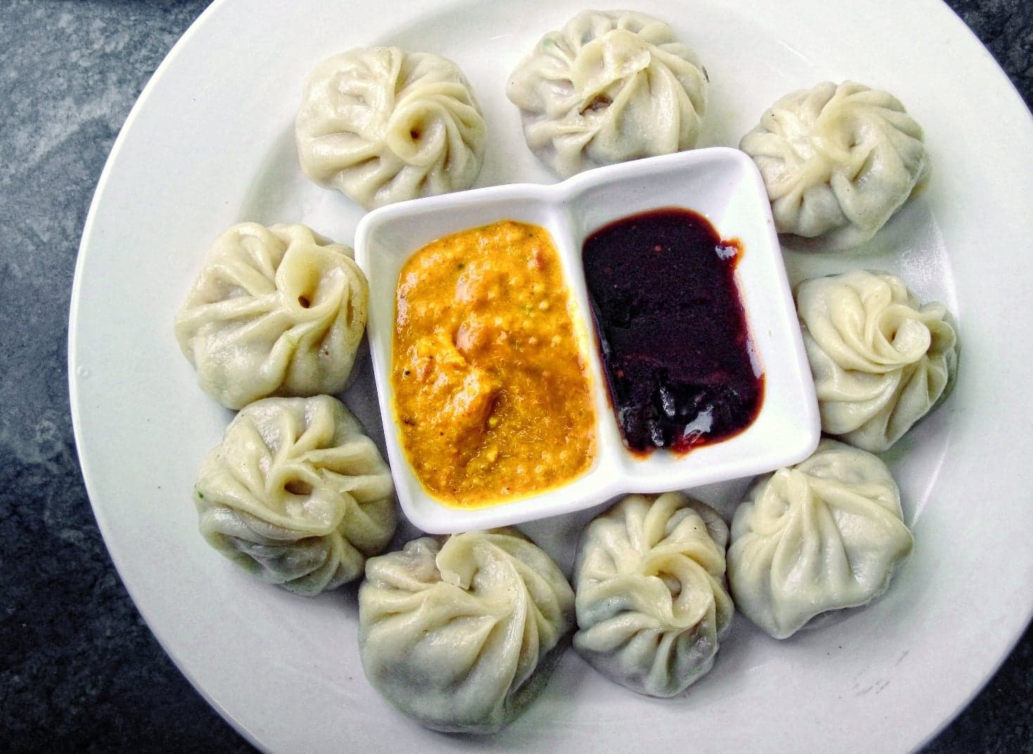 A bamboo steamer basket full of white momo dumplings with a dipping sauce beside them