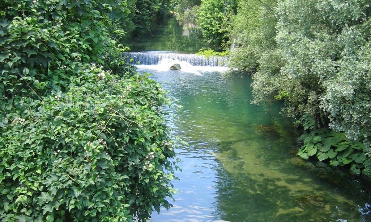Old wooden watermills perched on a weir above the Pliva River with autumn-coloured forest behind