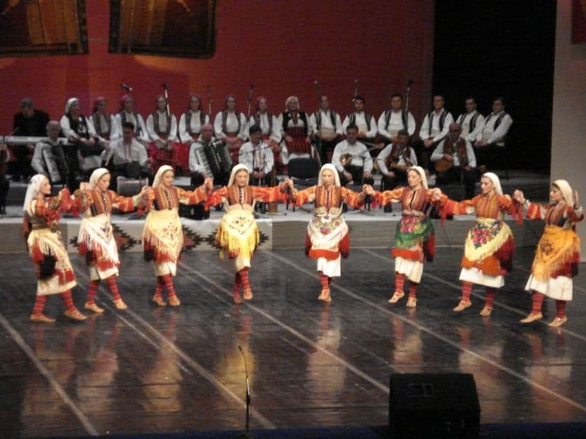 A large circle of Bulgarian folk dancers in colourful traditional costume holding hands and dancing outdoors