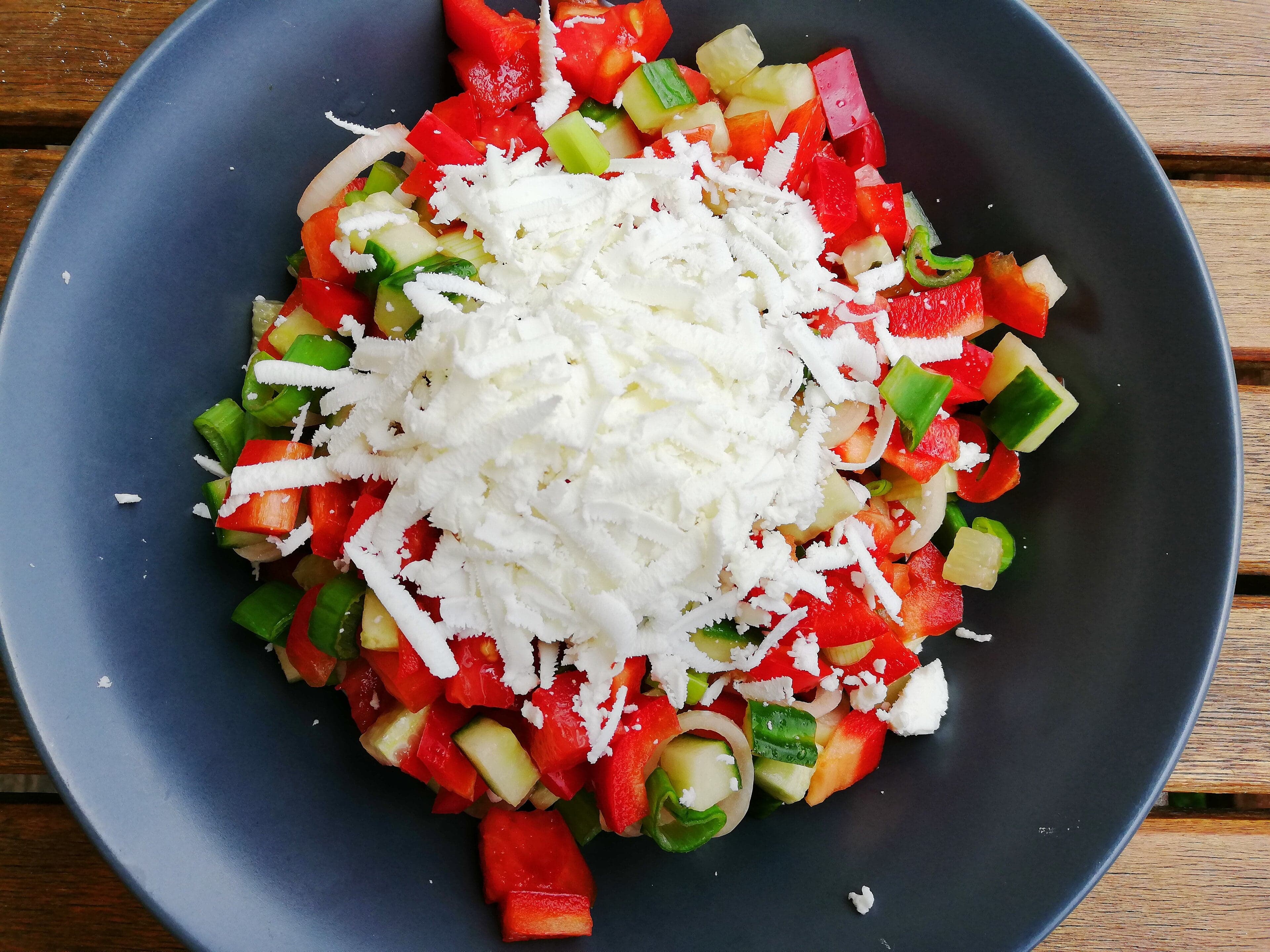 A bowl of shopska salad with red tomatoes, cucumbers, green peppers and a thick white layer of grated sirene cheese on top