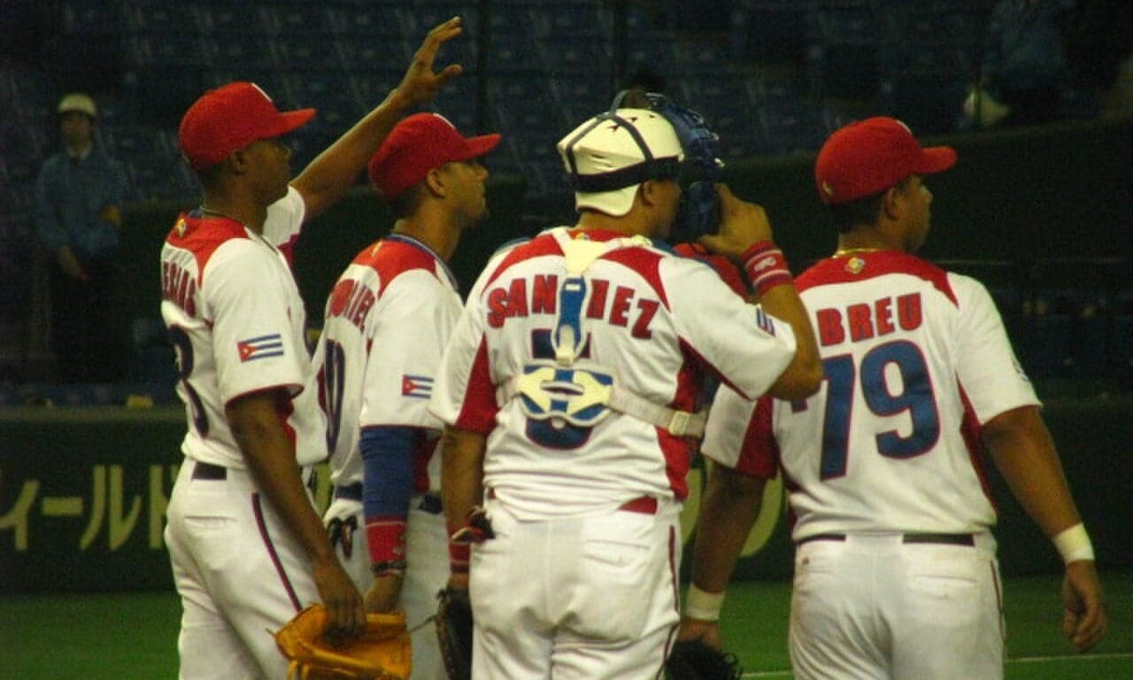A young Cuban baseball player in a white uniform at bat on a sunlit diamond