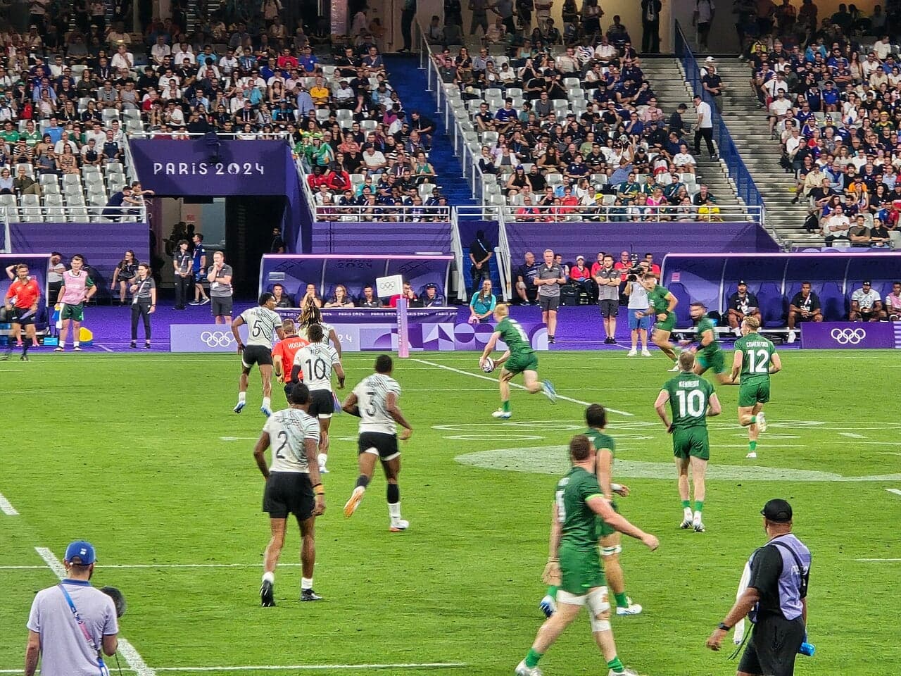 A Fiji rugby sevens player in white kit running with the ball against a blue background