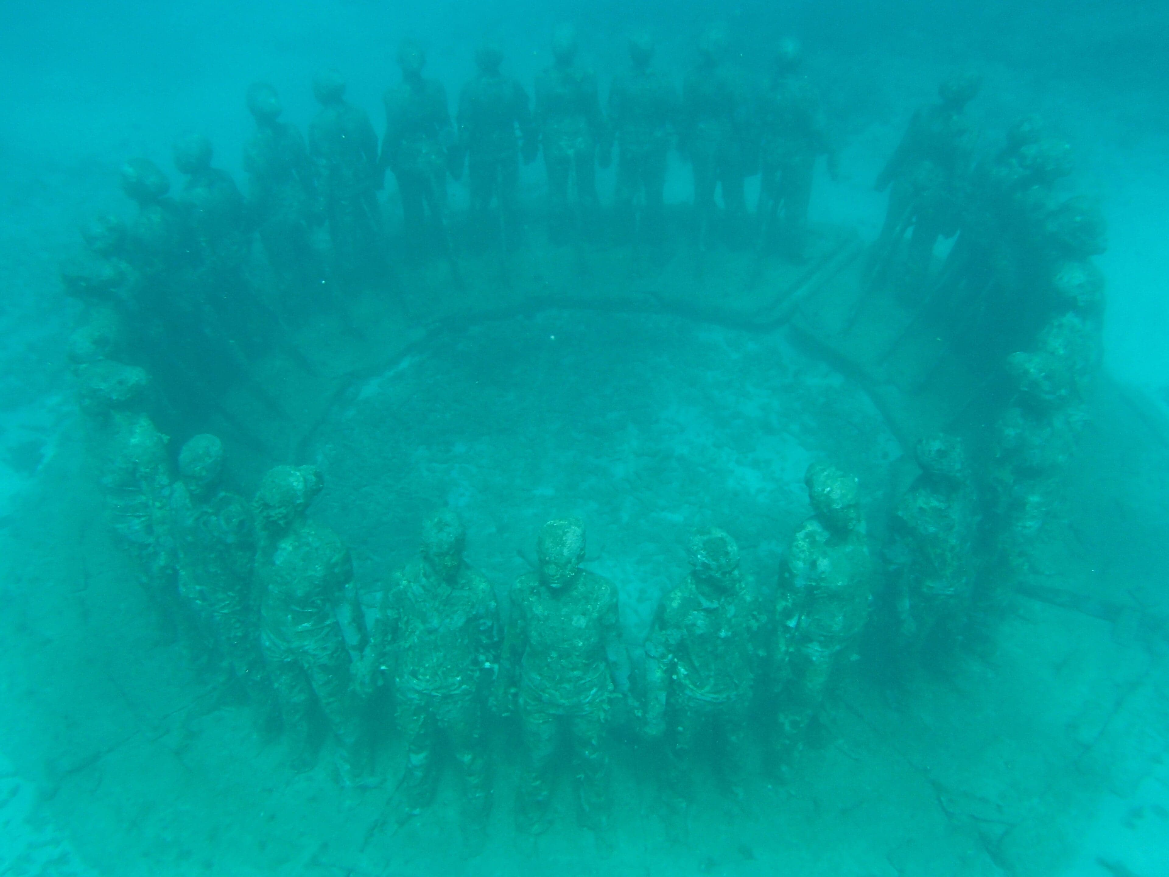 A circle of human-shaped sculptures on the seabed covered in coral and surrounded by tropical fish