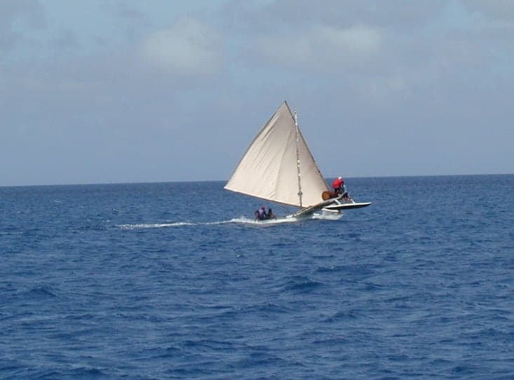 A carved wooden sailing canoe with a colourful sail crossing calm water between two Bijagós islands