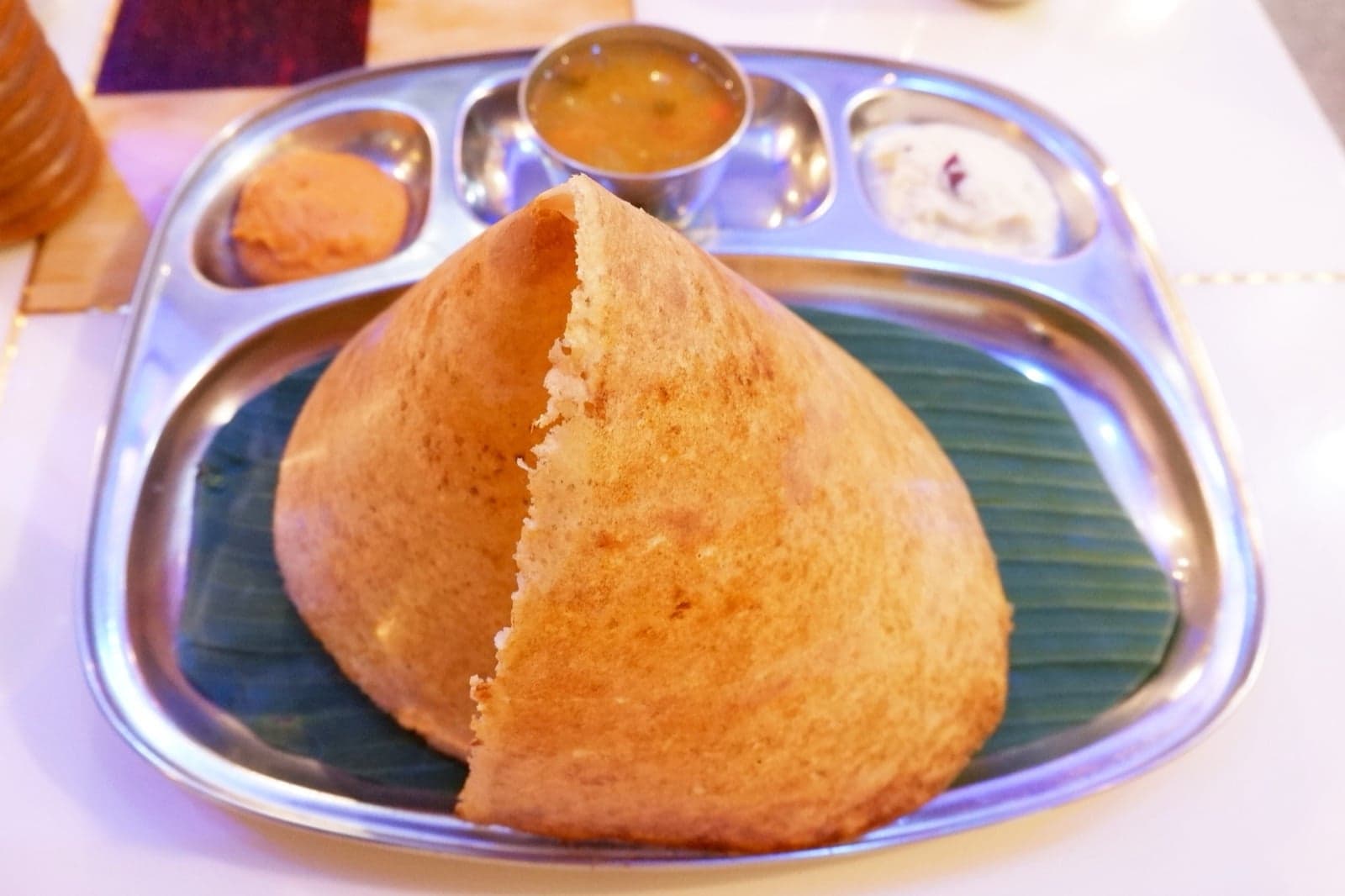 A cone-shaped golden dosa on a metal plate with small bowls of chutney and sambar