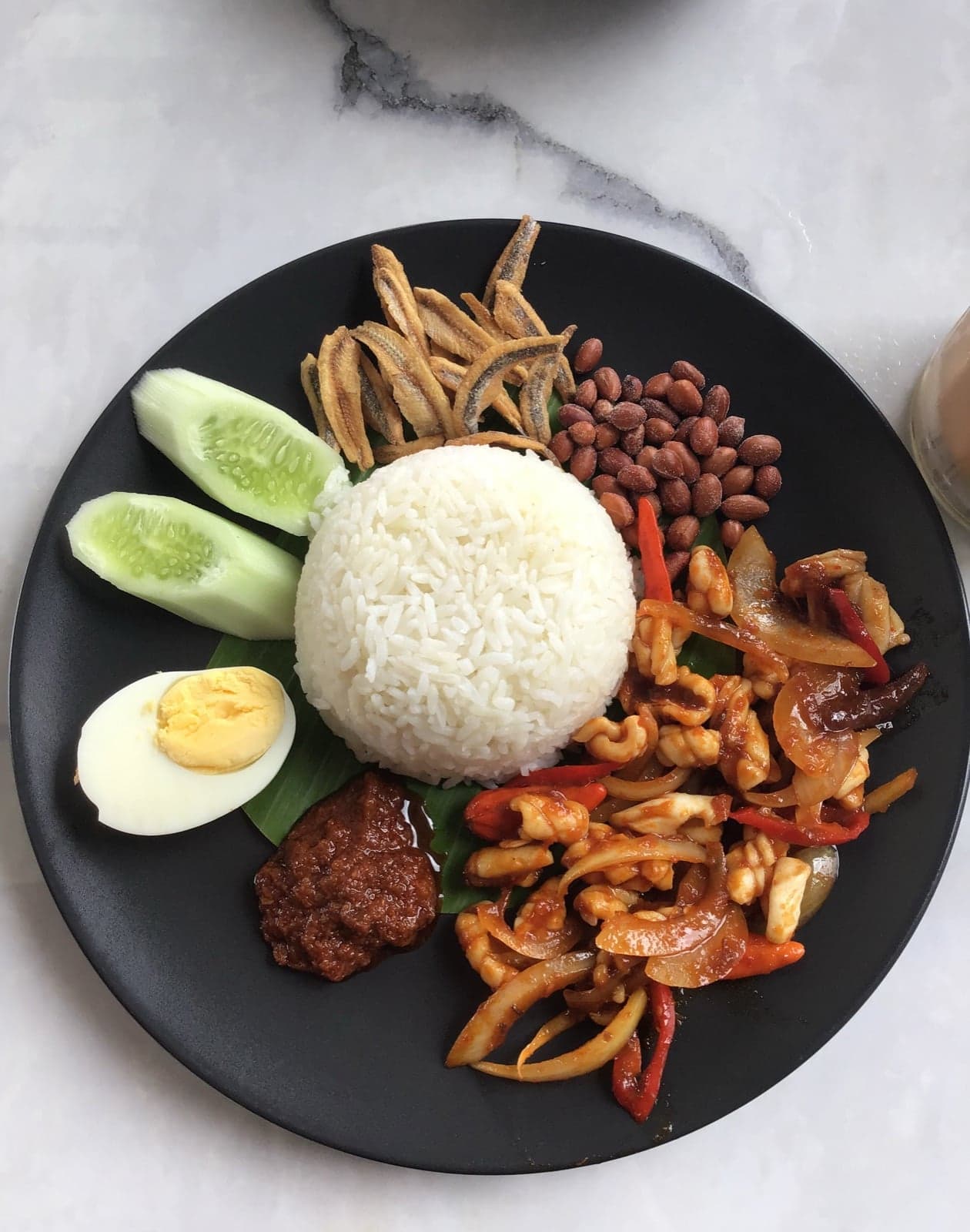 A plate of nasi lemak with coconut rice in the centre, surrounded by egg, peanuts, anchovies, cucumber and red sambal sauce