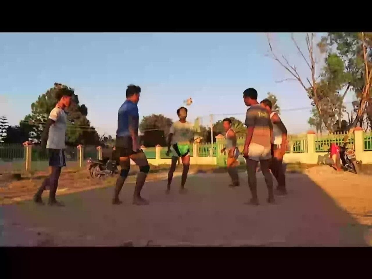 Six chinlone players in a circle, one kicking a woven cane ball into the air