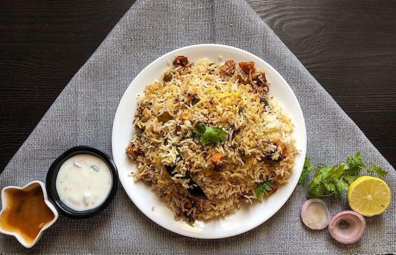 A plate of biryani - long-grain rice in shades of yellow, white and orange, with raita and lemon on the side
