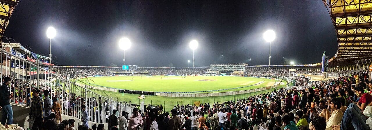 A packed cricket stadium at night, lit by floodlights, full of fans