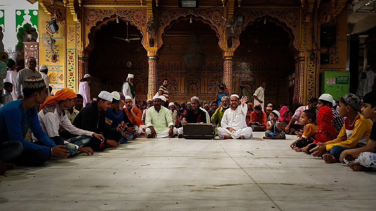 A group of qawwali singers and a harmonium player performing while a crowd sits around listening