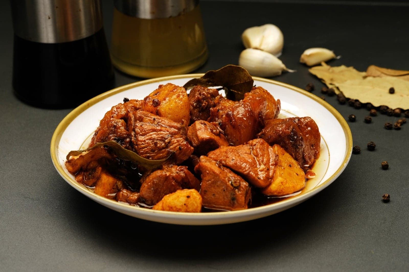 A white plate of adobo - dark, glossy chunks of meat with bay leaves and potatoes