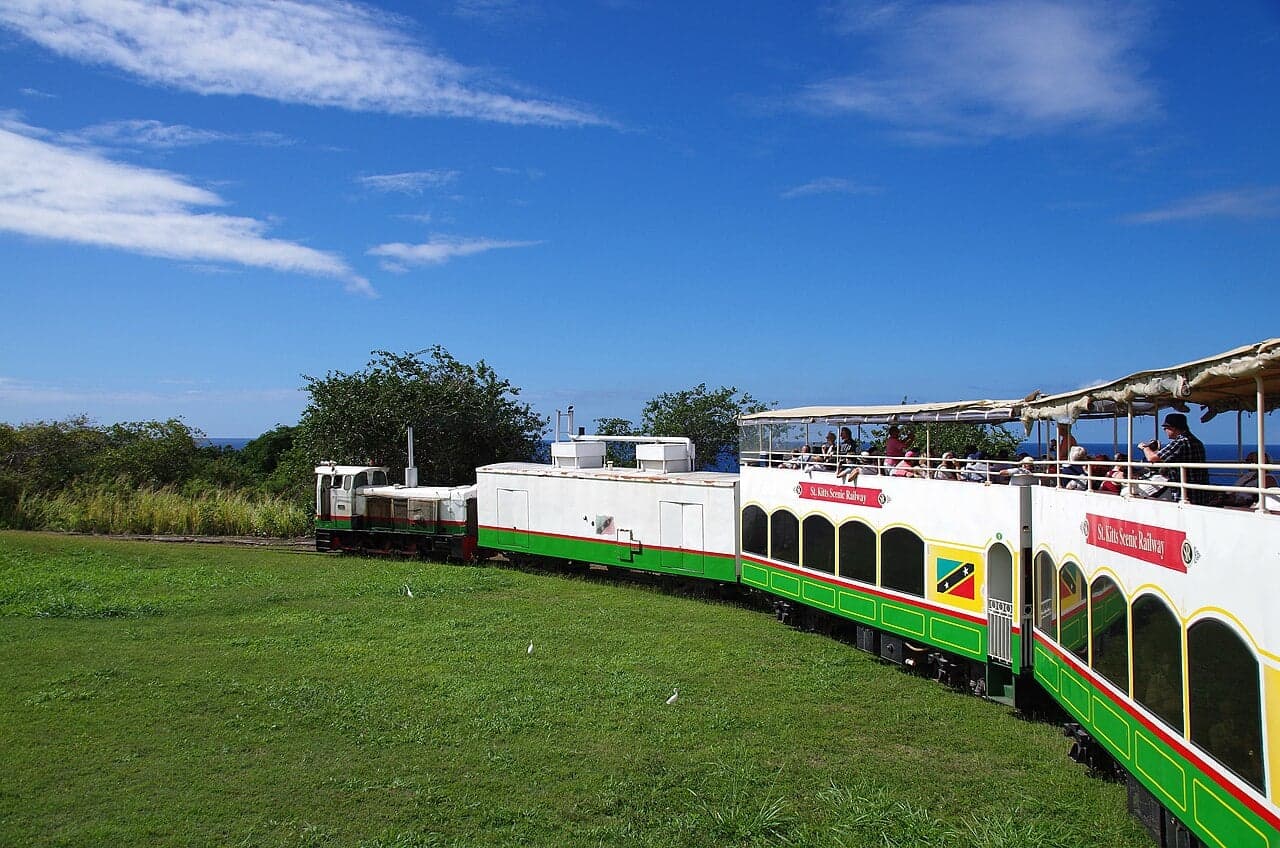 A small red and yellow train running through green countryside with the sea visible in the distance on Saint Kitts