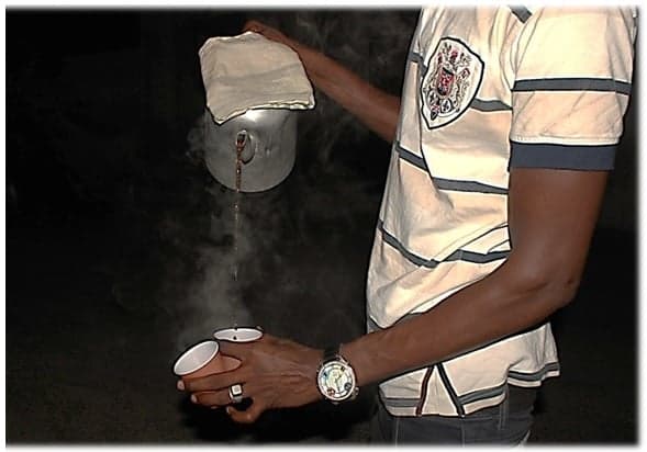 A small glass cup of Café Touba next to a metal pot