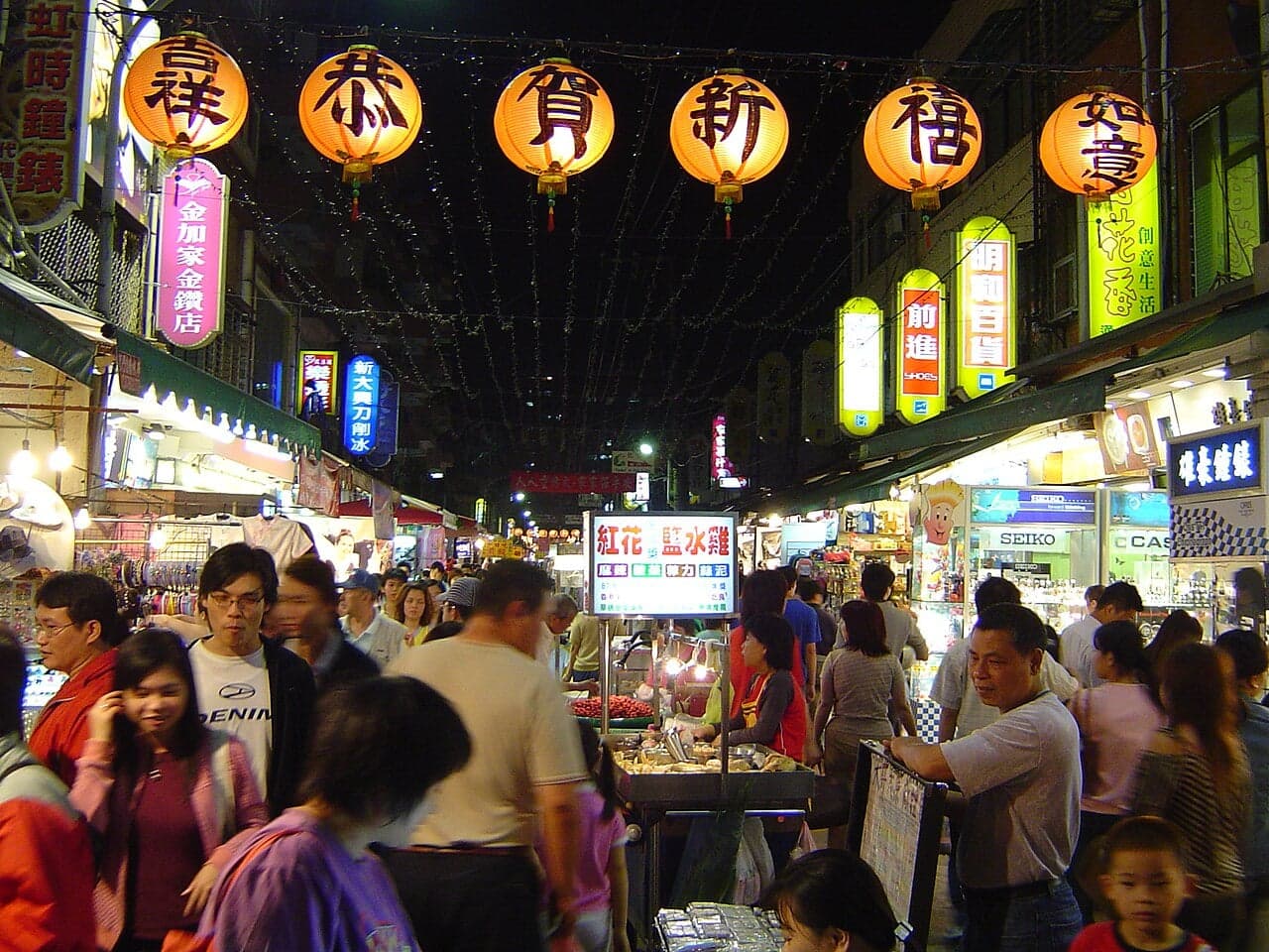 Crowds of people walking past brightly lit food stalls at a Taiwanese night market