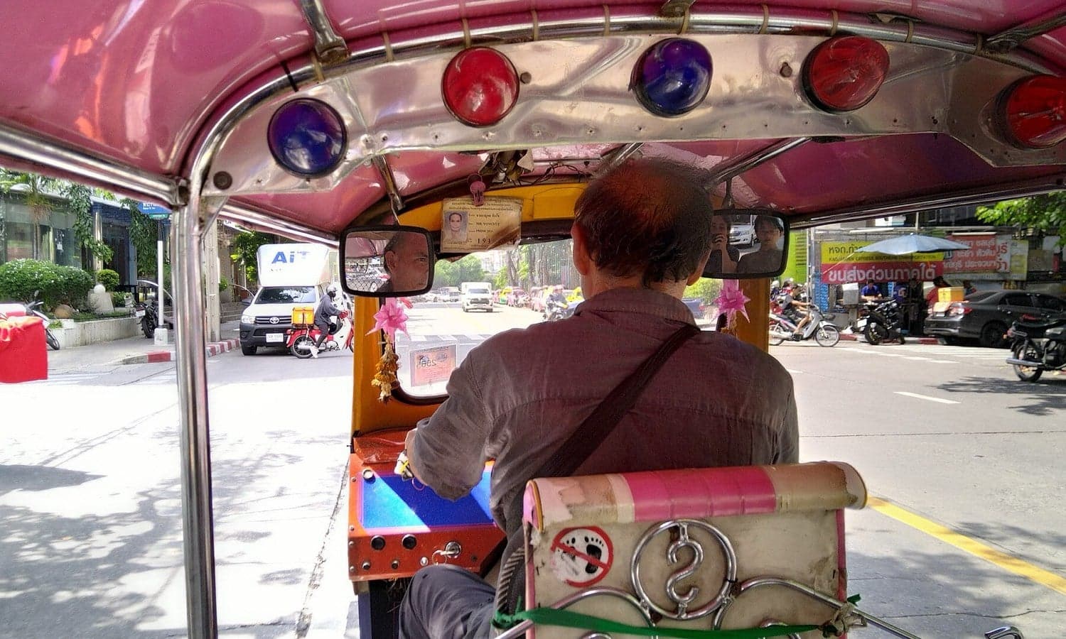View from inside a tuk-tuk in Bangkok, looking past the driver onto a sunny street