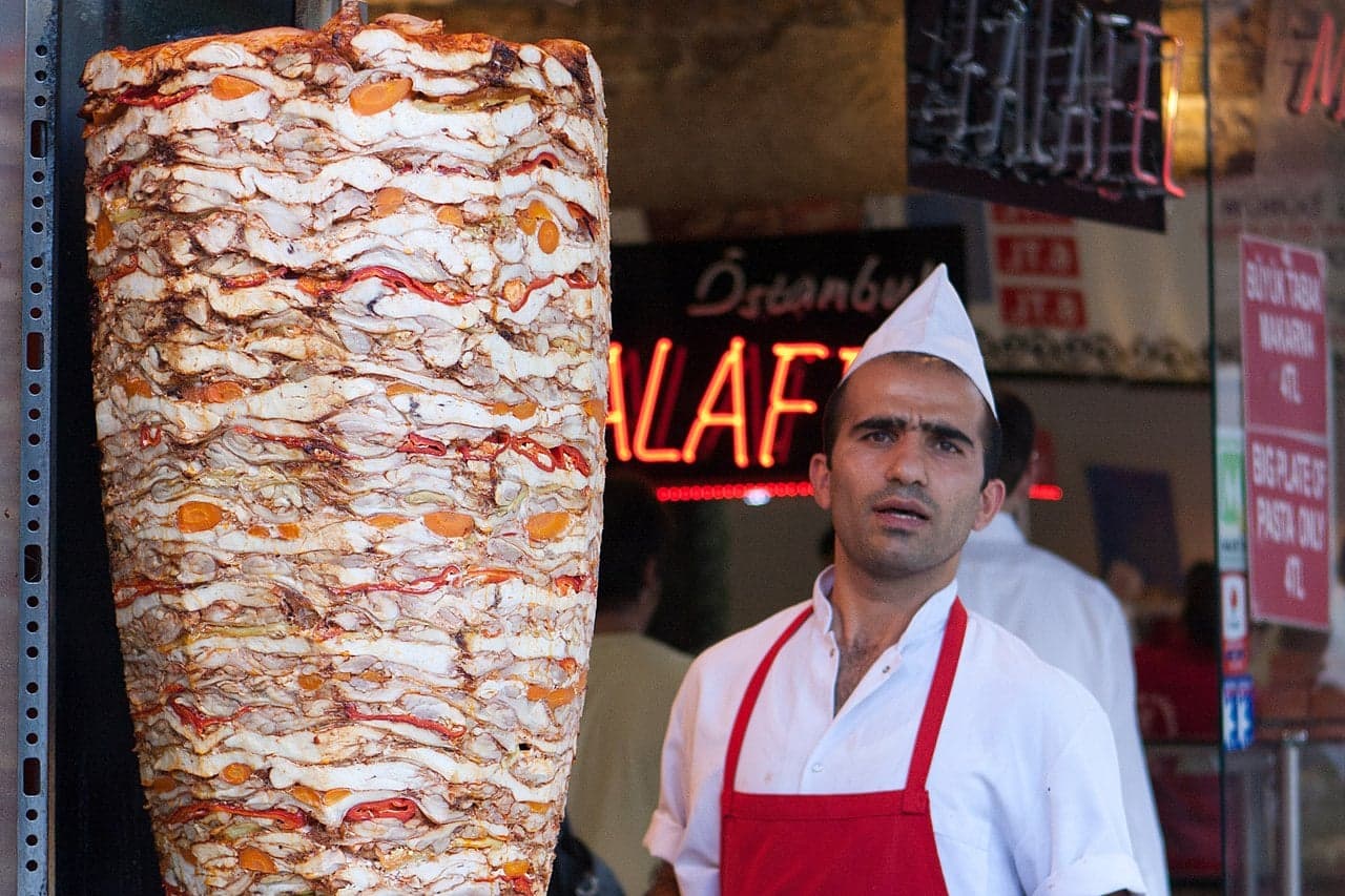 A tall vertical doner kebab spinning slowly in front of a flame in a Turkish kitchen
