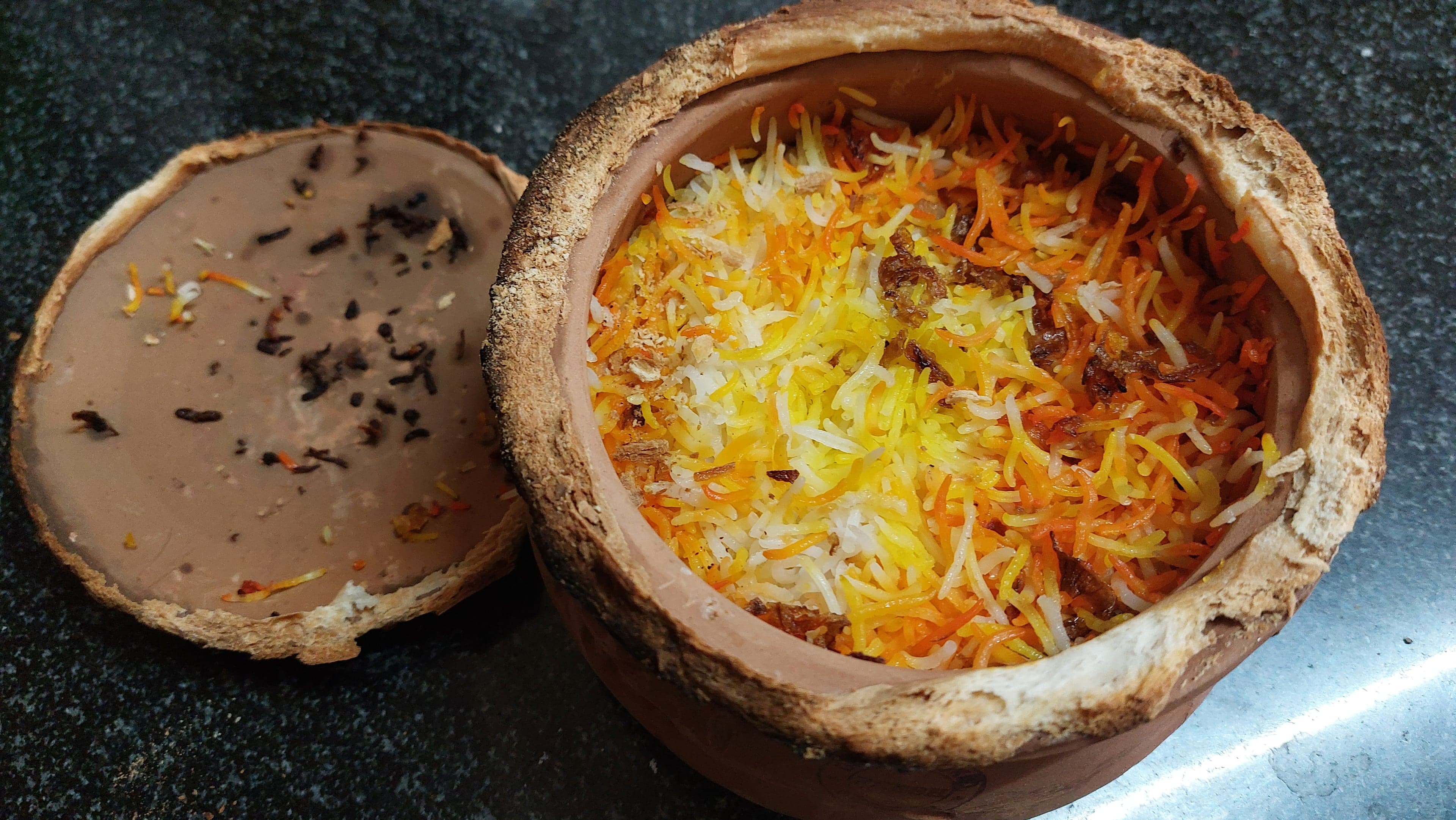 A clay pot full of bright yellow and orange biriyani rice, with the lid placed beside it