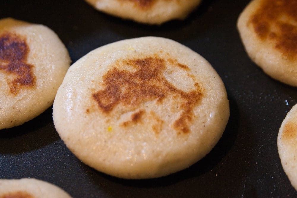 Several golden arepas cooking on a griddle, some with melted cheese