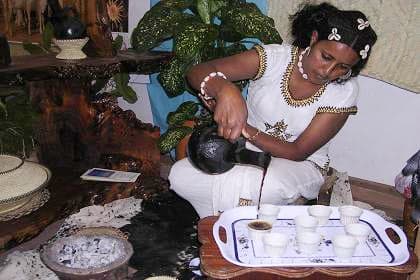 A woman in traditional dress pouring coffee from a clay jebena pot into small cups arranged on a tray