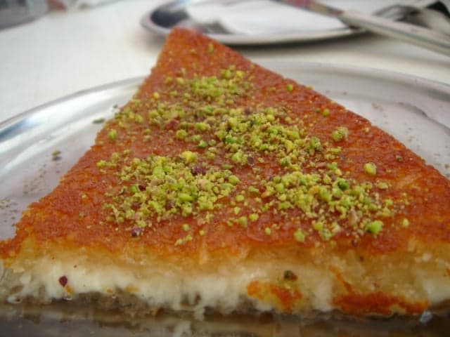 A round golden knafeh pastry sprinkled with crushed pistachios being cut into slices