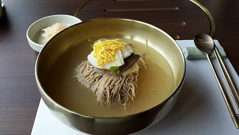 A bowl of naengmyeon cold noodles in clear broth topped with cucumber and a boiled egg