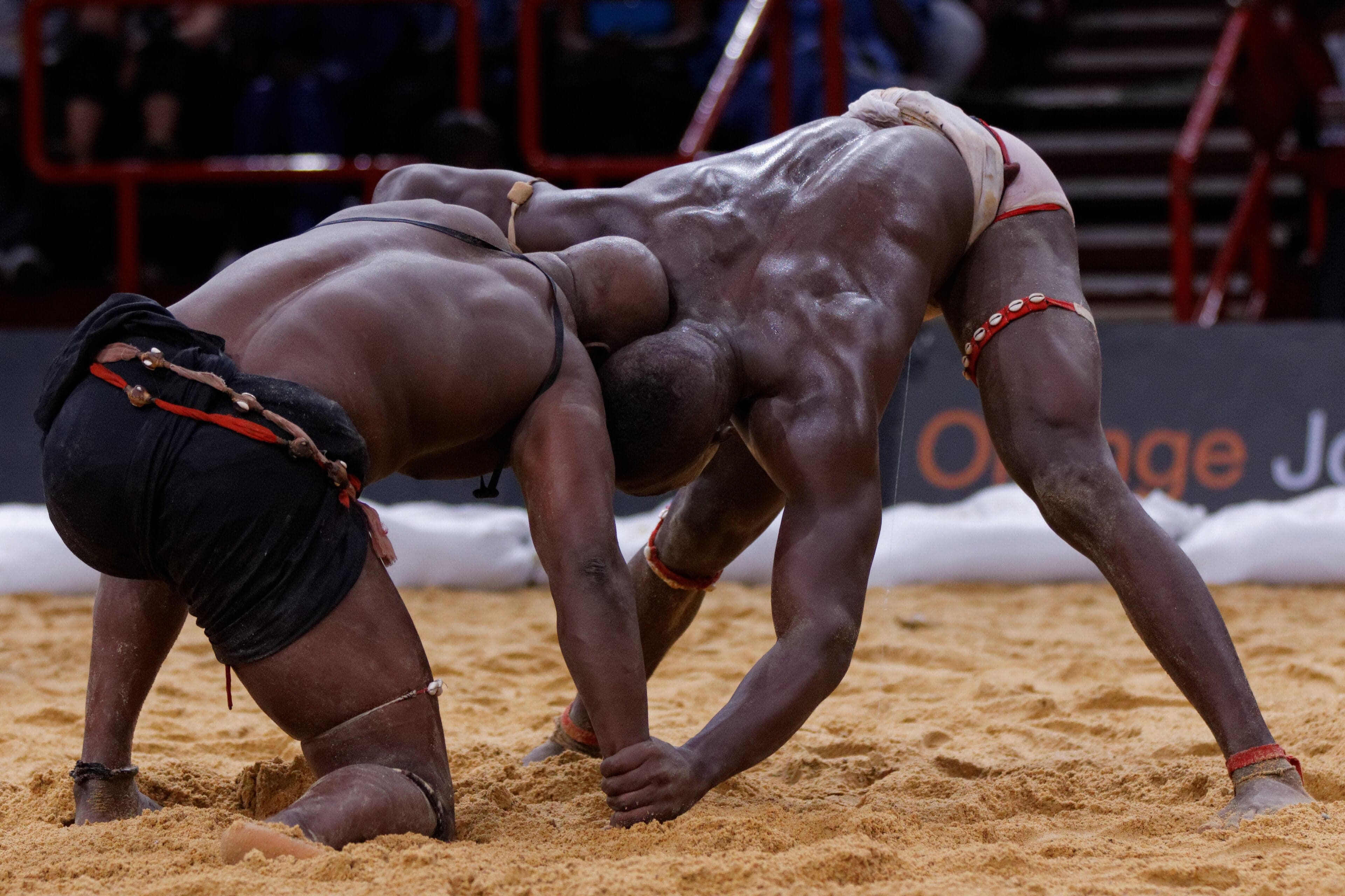 Two Senegalese wrestlers facing off in a sandy arena with a crowd watching